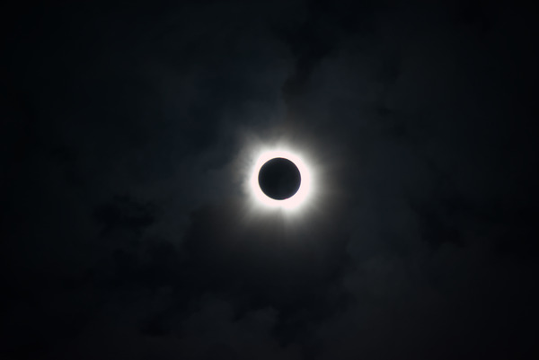 The sun, eclipsed by the
							moon. There is a bright glow around a black void. There is a hint
							of a pink burst on the lower-right side. The clouds surrounding
							the eclipse glow a dark purple.